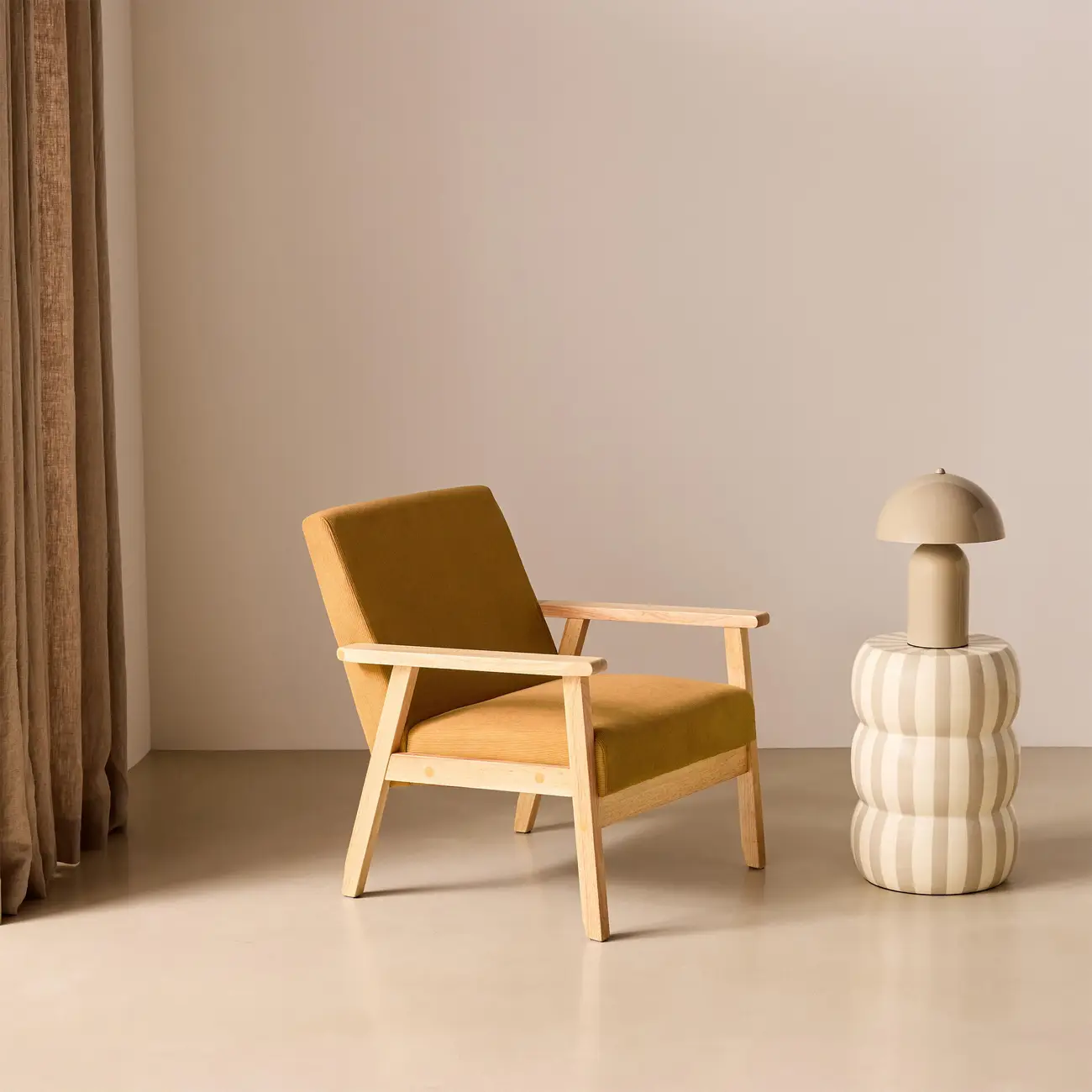Le même fauteuil moutarde mis en scène dans un studio beige, à côté d'un rideau en lin et d'une lampe sur un guéridon rayé